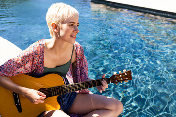 Naklejka premium Non-binary musician sitting poolside in sun, playing acoustic guitar near clear water, copy space