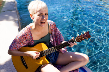 Non-binary person playing acoustic guitar at pool edge, sparkling water reflections, copy space