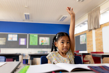 Young African American girl raising hand at desk in elementary school classroom with open workbook