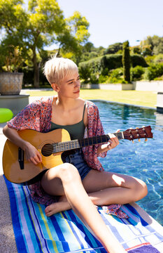 Non-binary guitarist sitting on striped pool towel by backyard pool edge, strumming acoustic guitar
