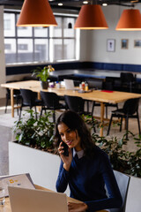 Middle Eastern woman juggling phone call and laptop in coworking space, with reports and plants