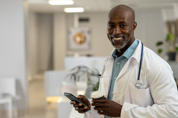 African American male doctor smiling, holding smartphone and coffee cup in clinic, copy space