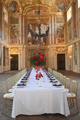 Generated ImageLuxurious wedding reception table setup with red roses centerpiece in a historic frescoed Italian palace, gold chairs, white linen, and navy napkins in elegant imperial style