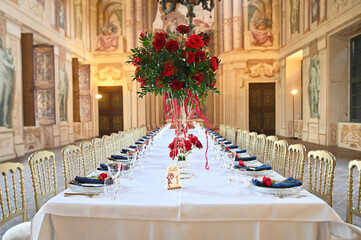 Romantic reception dinner setup with red roses, white tablecloths, golden chairs, and navy blue napkins in an opulent Italian baroque-style hall with classical frescoes