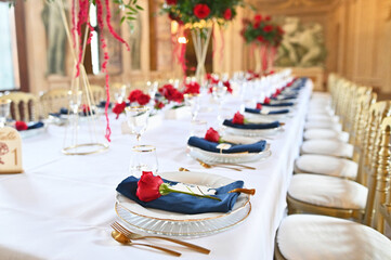 Close-up of refined wedding table decor with red rose on navy napkin, crystal plate and gold flatware for a luxurious romantic dinner setup in a frescoed palace hall
