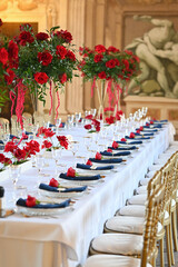 Luxurious wedding table setting with red roses, gold chairs, and navy napkins in a historic frescoed palace, elegant floral centerpieces and fine glassware ready for an upscale romantic celebration

