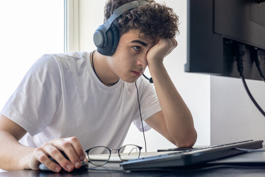 Sad and exhausted teenage boy with curly hair sitting at desk in front of computer monitor, experiencing digital fatigue and stress during distance learning. Concept of ADHD, cyberbullying, depression