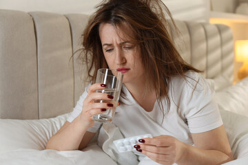 Unhappy woman with pills and glass of water suffering from hangover on bed at home