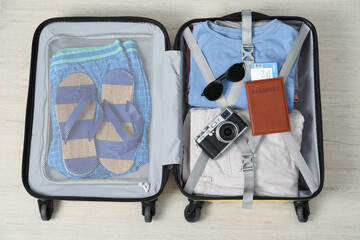 Different clothes, passport, camera and sunglasses in open suitcase on light wooden floor, top view