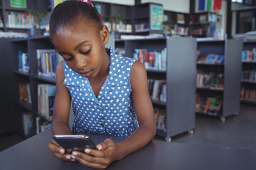 African American girl child using smartphone at library table, among bookshelves and book cart