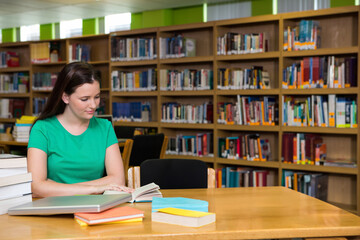 Woman reading open book at library study area, surrounded by laptop and book stacks, copy space