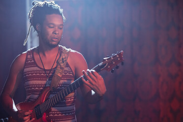 Male musician playing electric guitar on dim stage under colored lights, with decorated strap