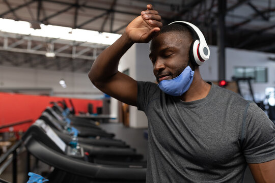 African American man wiping sweat on treadmill at gym with blue towel and mask, copy space