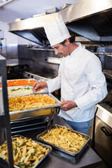 Male chef plating pasta and fries from buffet trays in commercial kitchen, with serving spoon