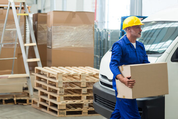 Middle-aged male worker carrying cardboard box in warehouse dock with pallets and van, copy space