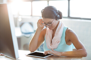 Obraz premium Woman sitting at desk in bright modern office, using tablet with headphones, glasses and pink scarf