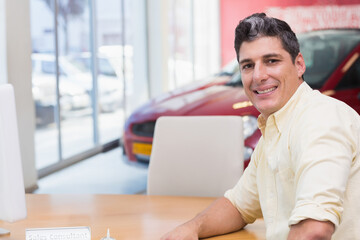 Man sitting at wooden desk in car dealership showroom with red display car, nameplate, copy space