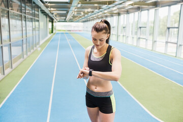 young adult woman checking smartwatch on blue track lanes in gym, wearing sports bra and shorts