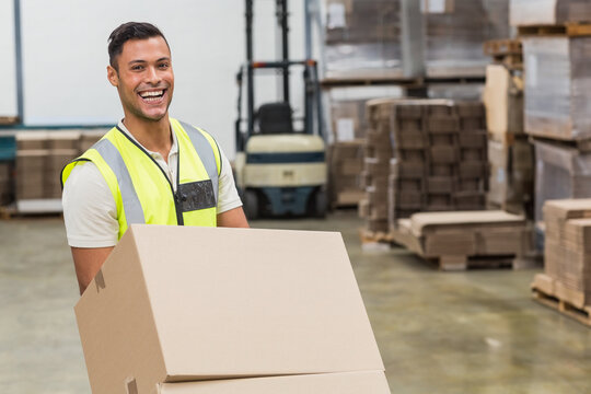 Man carrying stacked cardboard boxes in warehouse, with forklift and wooden pallets behind