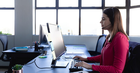 Young adult woman typing on keyboard at open-plan office desk, with headset and monitor, copy space