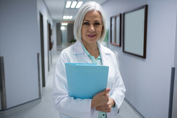 Female doctor standing under fluorescent lights in hospital corridor, holding blue medical folder