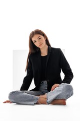 Young woman in a black blazer and jeans poses seated against a light background