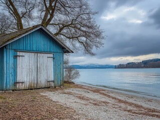 Nestled beside a serene lake an old wooden blue structure contrasts with white walls and a barren tree. The sandy beach reflects the gentle colors of a spring evening sky