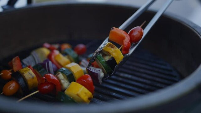 A person is cooking skewers of vegetables on a grill. The skewers are made of vegetables like zucchini, peppers, and onions. The person is using tongs to turn the skewers over on the grill