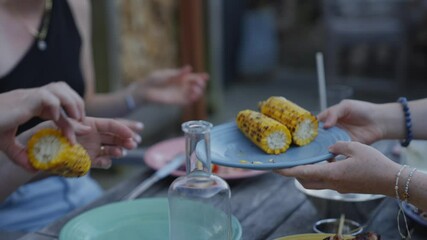 A group of people are eating corn on the cob. One person is holding a plate with corn on it and another person is holding a bottle. Scene is casual and relaxed