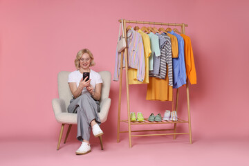 Smiling fashion stylist with smartphone and clothing rack on pink background