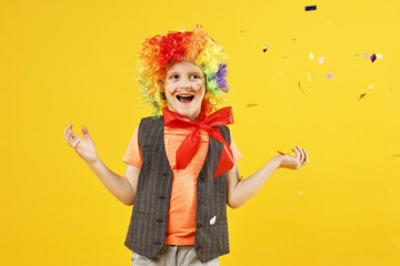 Happy little boy dressed like clown and flying confetti on orange background. Surprise party
