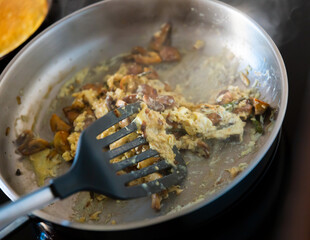 There is frying pan with scrambled eggs on stove. As flavor additive, pieces of champignons and green asparagus shoots were placed in dish.