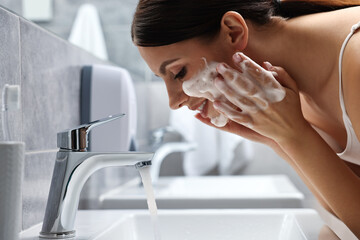 Woman washing her face with cleansing foam over sink in bathroom
