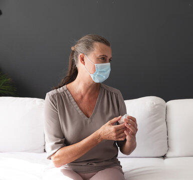 Senior woman wearing surgical mask holding white bottle sitting on sofa in living room
