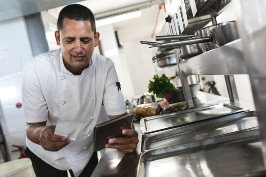 Male chef leaning over steel counter in commercial kitchen, reviewing ticket on tablet, copy space