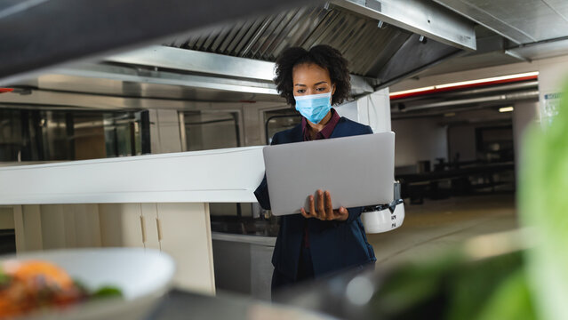 African American woman standing in kitchen under vent hood, examining laptop and robotic arm
