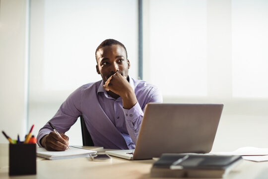 African American man writing at office desk in bright modern workspace, with laptop and notebook