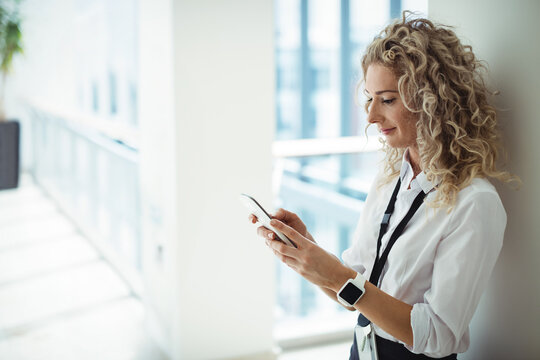 woman leaning in office hallway beside glass windows using smartphone with smartwatch, copy space