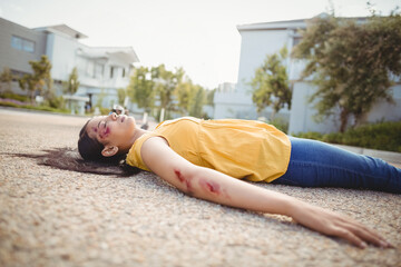 Woman lying flat on paved suburban street, with visible scrapes and nearby houses trees shrubs