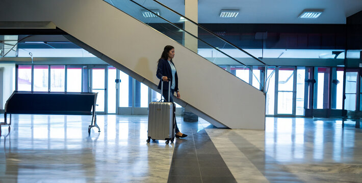 Mixed?Race young adult woman walking with silver suitcase at modern airport terminal near escalator