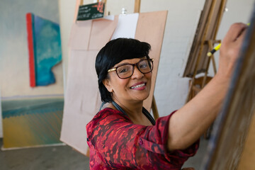 Mature Indian woman painting at wooden easel in art studio, with canvases and creative focus