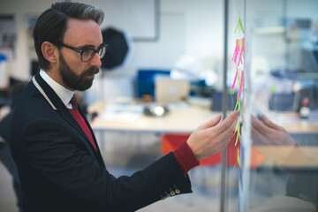 Asian man placing sticky notes on glass partition whiteboard in modern open-plan office, copy space