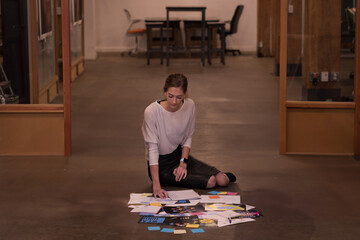 female designer kneeling on concrete floor in open-plan office, sorting papers and color swatches