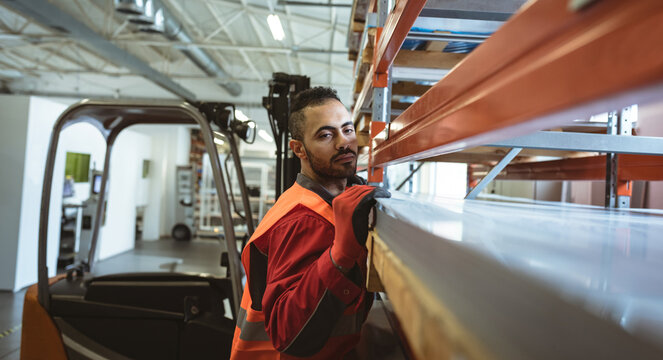 Male warehouse worker leaning against shelving racks in industrial warehouse, with forklift truck