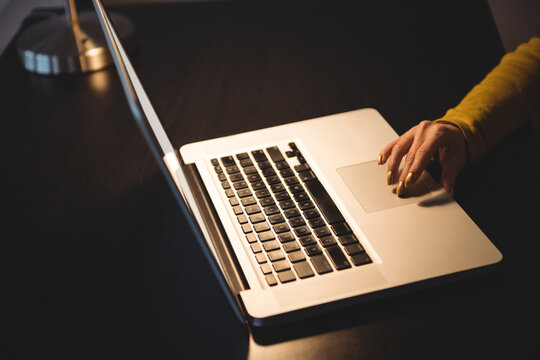 Right hand pressing trackpad on silver laptop at dark wooden office desk, under warm lamp glow
