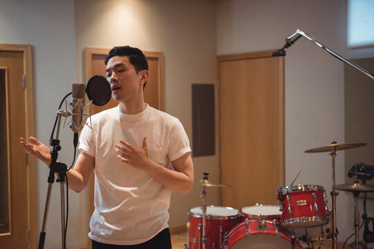 Young adult Asian man singing into condenser microphone in recording studio, with red drum kit
