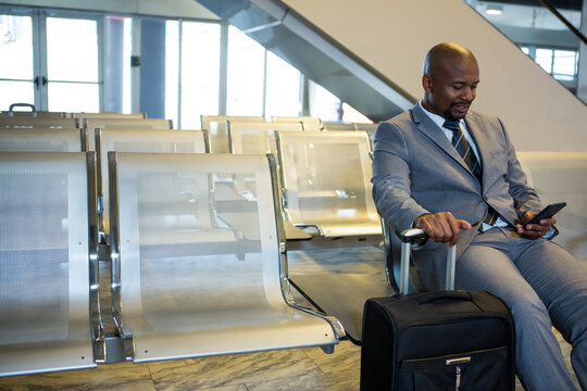 Mid adult African American man using smartphone on metal airport seating with suitcase, copy space