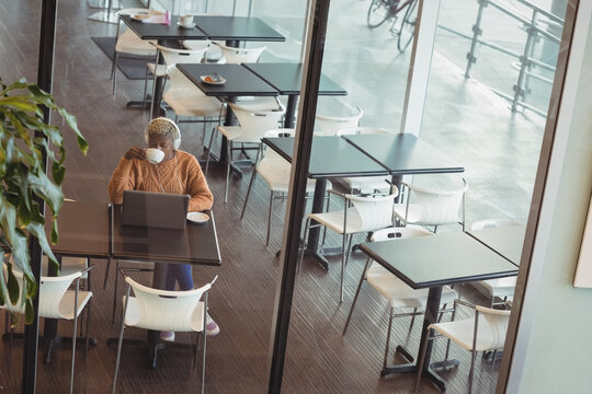 African American woman wearing headphones, using laptop, sipping coffee in cafe lounge, copy space