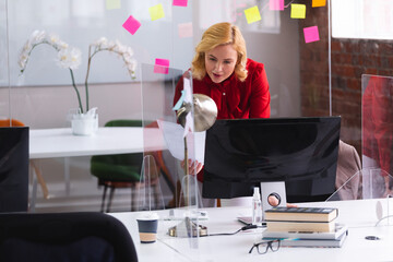 Professional woman leaning over desk with divider in open office, reviewing documents and monitor