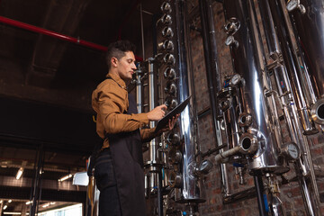 Middle-aged man operating tablet and monitoring stainless steel columns in distillery, copy space